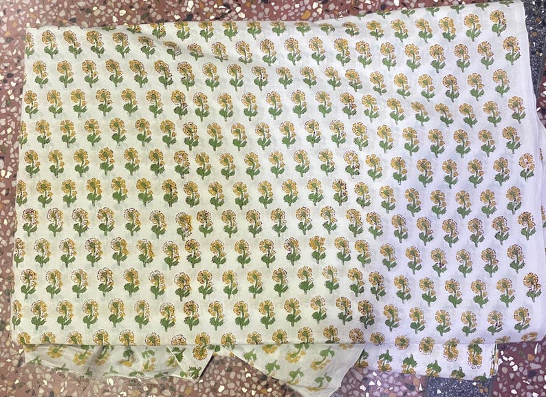 Op de afbeelding: Een gevouwen stuk witte stof met een herhalend patroon van kleine gele bloemen met groene stengels en bladeren. De bloemenprint is gerangschikt in consistente horizontale en verticale rijen over het gehele oppervlak van het textiel.