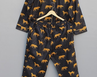 Puede incluir: Conjunto de pijama negro con estampado de leopardo dorado repetido. El conjunto incluye una camisa con botones y ribetes dorados y pantalones a juego con cintura elástica. El pijama está colgado de una percha de madera.