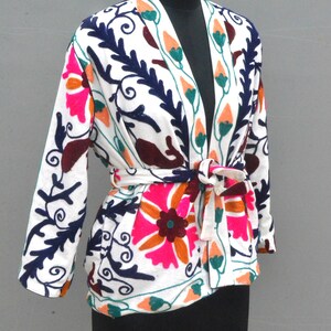 May include: White embroidered jacket with a floral pattern in shades of pink, orange, green, blue, and brown. The jacket has a tie belt and a V-neck.