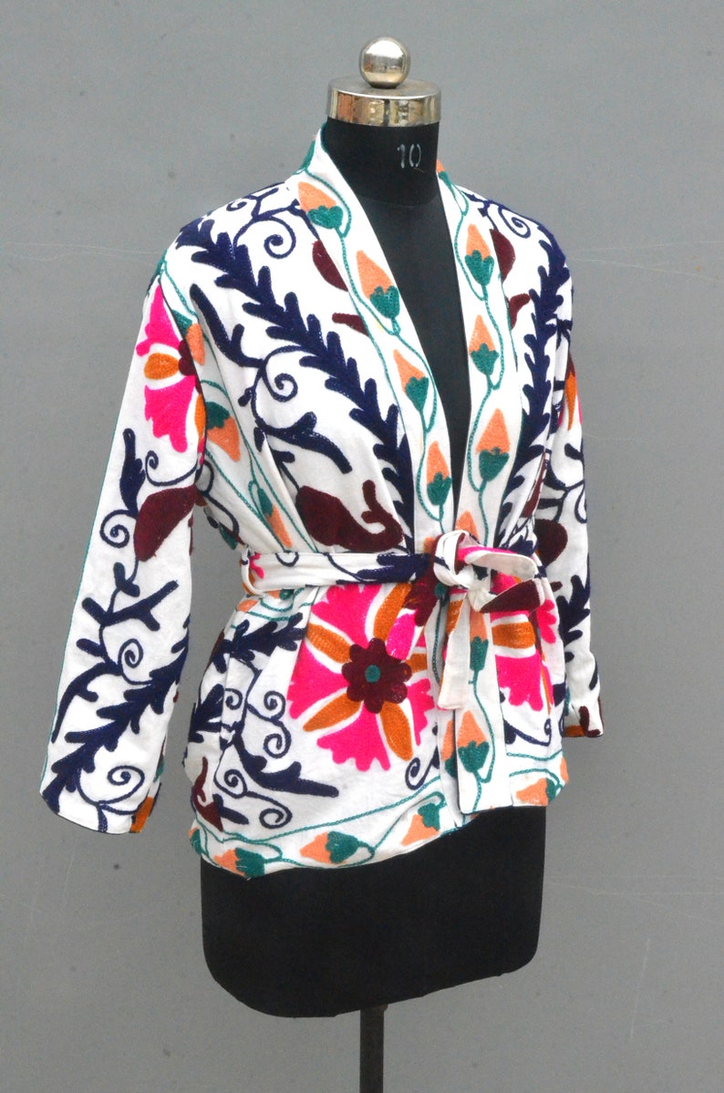 May include: White embroidered jacket with a floral pattern in shades of pink, orange, green, blue, and brown. The jacket has a tie belt and a V-neck.