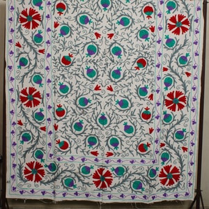 May include: White fabric with a floral embroidery pattern. The design features red, green, and purple flowers with gray stems and leaves. The fabric has a white border with a purple and green trim.