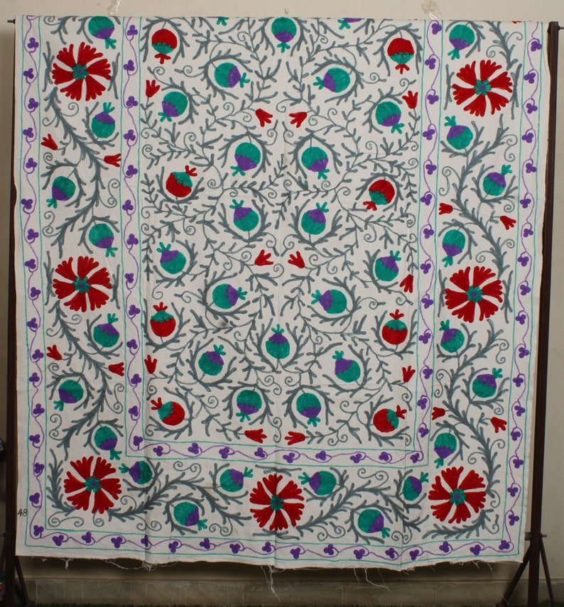 May include: White fabric with a floral embroidery pattern. The design features red, green, and purple flowers with gray stems and leaves. The fabric has a white border with a purple and green trim.