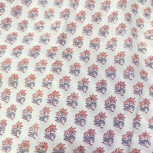 Op de afbeelding: Een witte stof met een herhalend blokdrukpatroon van kleine bloemmotieven. Elke individuele bloem bestaat uit zacht perzikkleurige bloemblaadjes gecombineerd met gedempte blauwgrijze bladeren en stengels in een regelmatig raster.