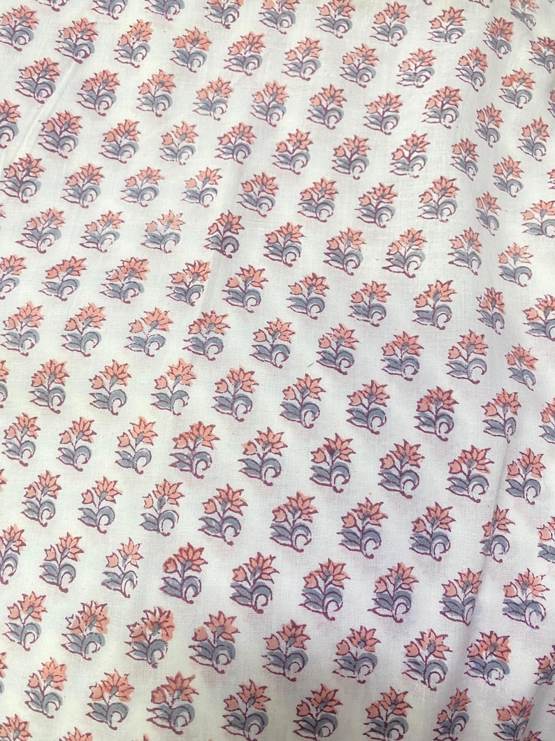 Op de afbeelding: Een witte stof met een herhalend blokdrukpatroon van kleine bloemmotieven. Elke individuele bloem bestaat uit zacht perzikkleurige bloemblaadjes gecombineerd met gedempte blauwgrijze bladeren en stengels in een regelmatig raster.