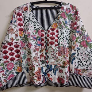 May include: A quilted jacket with a floral patchwork design. The jacket features a V-neck, long sleeves, and a button-up front. The floral patterns include pink, red, blue, and yellow flowers on a white background. The cuffs and inner lining have black and white stripes.