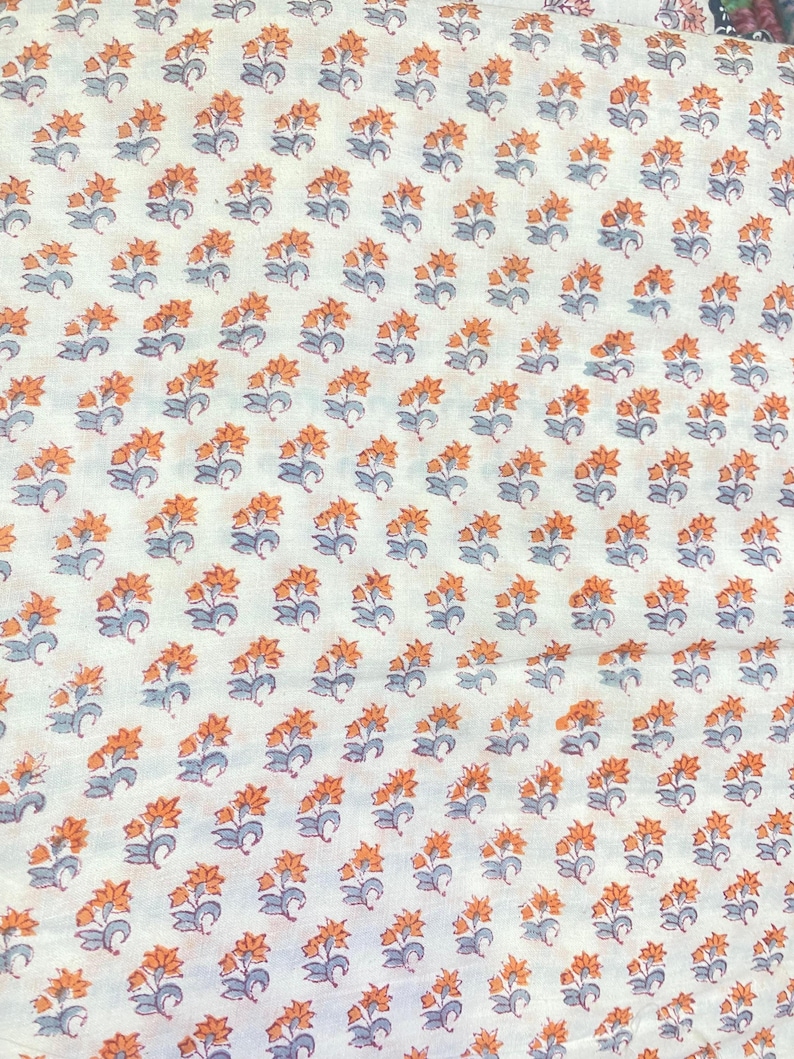 Op de afbeelding: Een witte stof met een herhalend patroon van kleine oranje bloemen met grijsblauwe stengels en bladeren. Het bloemmotief is in een consistent rasterachtig patroon over het gehele oppervlak van het textielmateriaal gerangschikt.