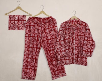 May include: A red and white floral print pajama set, including a long-sleeved button-down shirt, pants, and a matching eye mask, all hanging on wooden hangers. The pajamas have white trim.