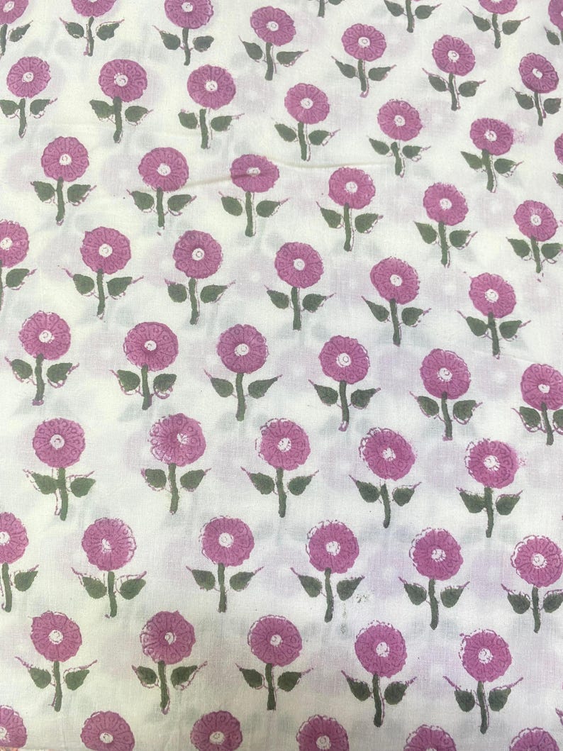 Op de afbeelding: Een witte stof met een herhalend patroon van kleine roze bloemen met donkergroene stengels en bladeren. Het bloemmotief is in een rasterachtig patroon over het oppervlak verdeeld, wat een consistente botanische print op het textiel laat zien.