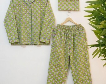 May include: A pyjama set consisting of a long-sleeved button-down shirt and matching trousers, accompanied by a small envelope-style pouch. The fabric is light green with a repeating floral pattern in pink and white, featuring dark green piping.