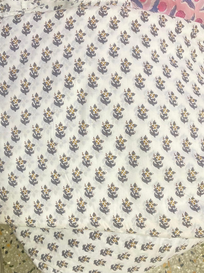 Op de afbeelding: Een witte stof met een herhalend patroon van kleine, gestileerde bloemen in gedempt grijs met gele centra. De bloemenprint is in een consistent rasterpatroon over het textiel verdeeld, wat zorgt voor een uniform en repetitief visueel ontwerp.