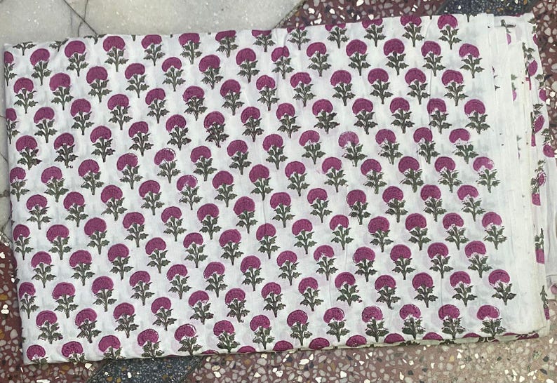 Op de afbeelding: Een gevouwen stuk witte stof met een herhalend blokdrukpatroon van kleine, donkergroene stengels en bladeren, bekroond met afgeronde, levendige magentakleurige bloemhoofdjes die in een raster over het hele textiel zijn verdeeld.