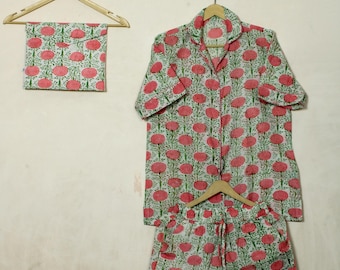 Op de afbeelding: Een driedelige pyjamaset met een overhemd met knopen, een korte broek en een bijpassende etui, allemaal versierd met een herhalend patroon van roze bloemmotieven op een witte achtergrond. De set wordt gepresenteerd op houten hangers tegen een neutrale achtergrond.