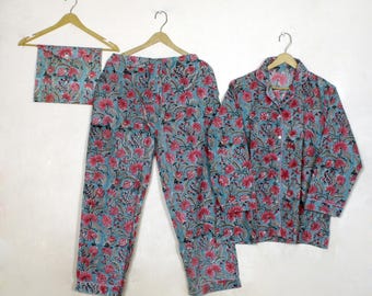 以下が含まれることがあります： ピンクとグレーの花柄の、水色のパジャマセット。長袖のボタンダウンシャツ、パンツ、お揃いのアイマスクが含まれており、すべて木製のハンガーに掛けられています。