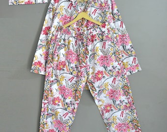 May include: White pajama set with a vibrant floral pattern in pink, yellow, and green. The set includes a long-sleeved button-down shirt, pants, and a matching pouch. The pajamas are displayed on a wooden hanger against a neutral background.