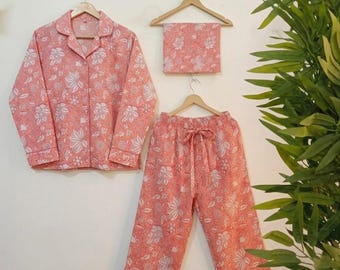 May include: A coral-colored pajama set with a white floral pattern. The set includes a button-down shirt, pants with a drawstring waist, and a matching eye mask. The pajamas are hanging on wooden hangers against a white wall.