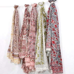 Könnte beinhalten: Fünf bunte bedruckte Schals hängen an einer weißen Wand. Die Schals sind aus Baumwolle und haben verschiedene florale und geometrische Muster. Die Schals sind in den Farben Rosa, Rot, Grün und Braun.
