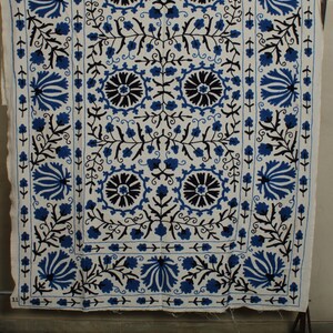 May include: A white fabric wall hanging with a blue and black floral embroidery design. The design features a repeating pattern of stylized flowers and leaves.