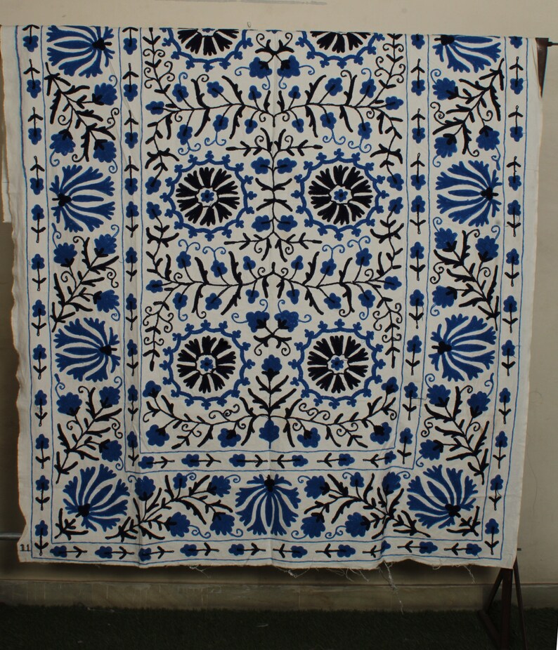 May include: A white fabric wall hanging with a blue and black floral embroidery design. The design features a repeating pattern of stylized flowers and leaves.