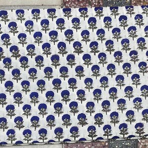Op de afbeelding: Een rechthoekig stuk witte stof met een herhalend patroon van kleine, donkerblauwe cirkelvormige bloemmotieven met groene stengels en bladeren. Het textiel is gevouwen en toont een consistente, gelijkmatig verdeelde botanische print.