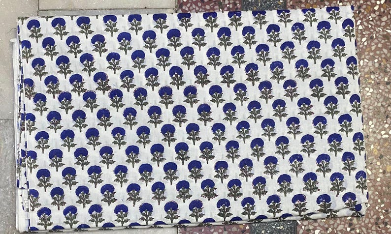 Op de afbeelding: Een rechthoekig stuk witte stof met een herhalend patroon van kleine, donkerblauwe cirkelvormige bloemmotieven met groene stengels en bladeren. Het textiel is gevouwen en toont een consistente, gelijkmatig verdeelde botanische print.