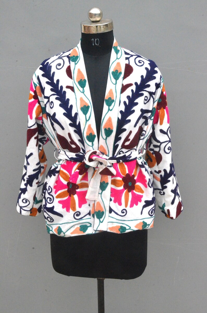 May include: White cotton jacket with a floral embroidery design in pink, orange, green, and blue. The jacket has a tie belt and a V-neck.