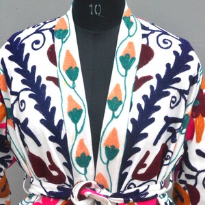 May include: White cotton robe with colorful floral embroidery. The robe has a long sleeve and a tie belt. The embroidery features flowers, leaves, and vines in shades of pink, orange, green, blue, and burgundy.