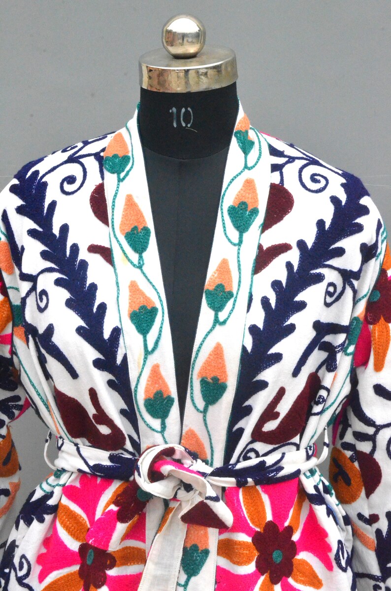 May include: White cotton robe with colorful floral embroidery. The robe has a long sleeve and a tie belt. The embroidery features flowers, leaves, and vines in shades of pink, orange, green, blue, and burgundy.