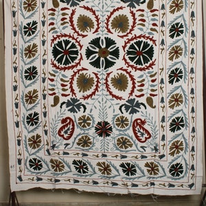 May include: White cotton fabric with intricate embroidery in shades of red, green, blue, and brown. The design features a central floral motif surrounded by smaller floral patterns and paisley shapes. The fabric has a border of repeating floral designs.