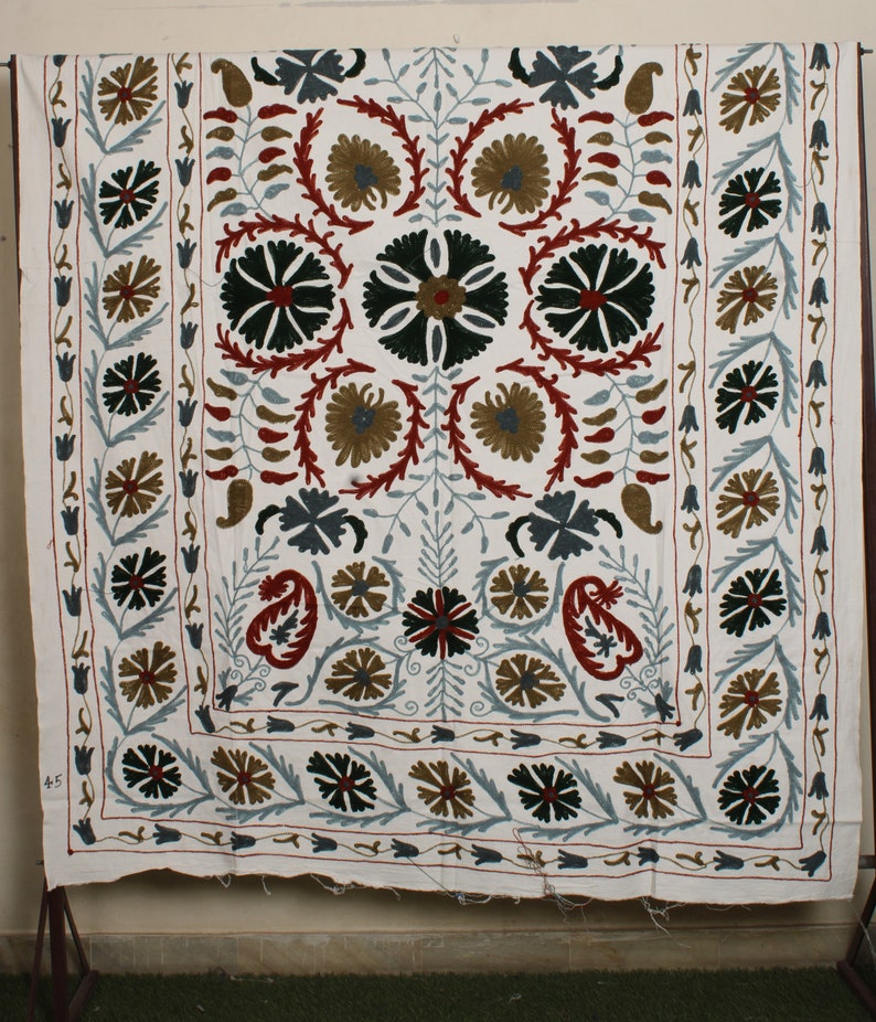 May include: White cotton fabric with intricate embroidery in shades of red, green, blue, and brown. The design features a central floral motif surrounded by smaller floral patterns and paisley shapes. The fabric has a border of repeating floral designs.