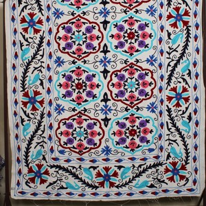 May include: A white cotton fabric with a colorful embroidered design. The design features a repeating pattern of floral motifs in shades of pink, blue, purple, and red. The fabric is edged with a black and blue border.