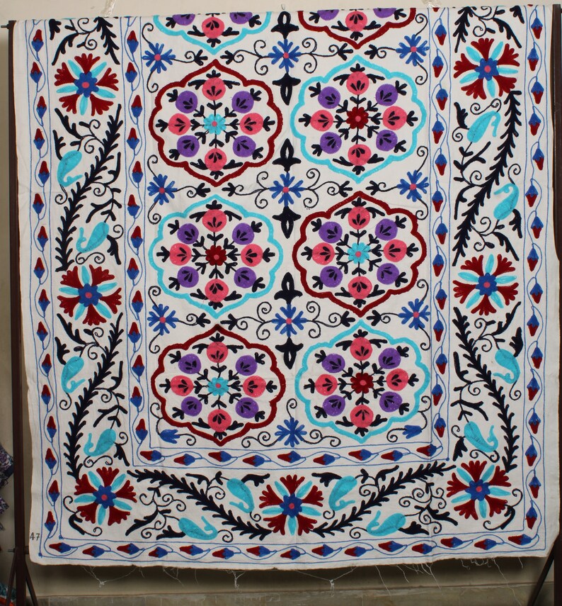 May include: A white cotton fabric with a colorful embroidered design. The design features a repeating pattern of floral motifs in shades of pink, blue, purple, and red. The fabric is edged with a black and blue border.