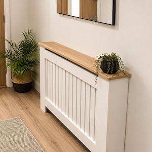May include: A white radiator cover featuring a vertical slat design and a light oak wood-toned top shelf. The cover is rectangular with a flat surface, providing a shelf for decorative items. The structure is clean-lined and functional.