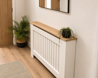 May include: A white radiator cover featuring a vertical slat design and a light oak wood-toned top shelf. The cover is rectangular with a flat surface, providing a shelf for decorative items. The structure is clean-lined and functional.