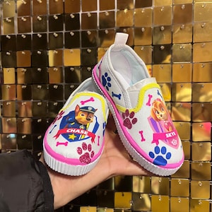 May include: Pair of white slip-on shoes featuring Paw Patrol characters Chase and Skye. The shoes have pink and yellow accents, paw prints, and bone designs. The soles are white with pink glitter trim.
