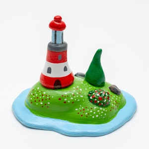May include: A hand-painted ceramic lighthouse figurine on a green island with a small tree and red flowers. The lighthouse is red and white with a gray roof and a blue window.