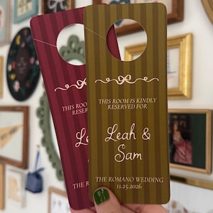May include: Two rectangular door hangers with a circular cutout at the top. One is dark red and the other is olive green, both featuring vertical stripes and a decorative bow graphic. Text reads: THIS ROOM IS KINDLY RESERVED FOR Leah & Sam THE ROMANO WEDDING 11.25.2026.