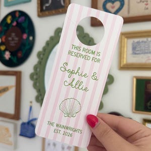 May include: A rectangular door hanger with a pink and white vertical striped pattern. The text reads THIS ROOM IS RESERVED FOR Sophie & Allie, with a green seashell illustration below and the text THE WAINWRIGHTS EST. 2026 at the bottom.
