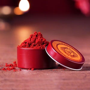 Puede incluir: Un recipiente de metal rojo con polvo rojo en su interior. El recipiente tiene una tapa con un diseño dorado de un símbolo.