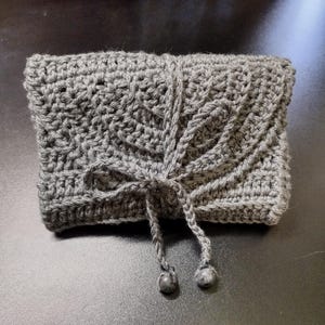 May include: A gray crocheted clutch with a textured pattern and a drawstring closure. The handmade bag is tied with a matching cord and features two small, decorative beads at the ends.