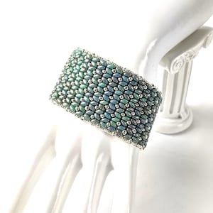 May include: Wide beaded bracelet in shades of teal and silver. The bracelet is made of small, oval-shaped beads arranged in a repeating pattern. The edges are finished with a silver border. The bracelet is displayed on a white, sculpted hand.