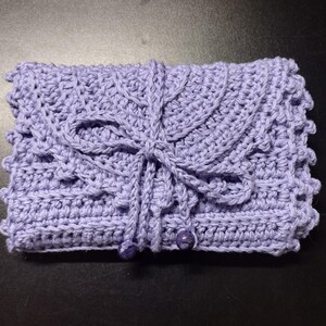 May include: A handmade, lavender crochet clutch. The clutch features a detailed, textured stitch pattern, a decorative flap, and a tie closure. Two small, round beads secure the closure. The overall design is intricate and delicate.
