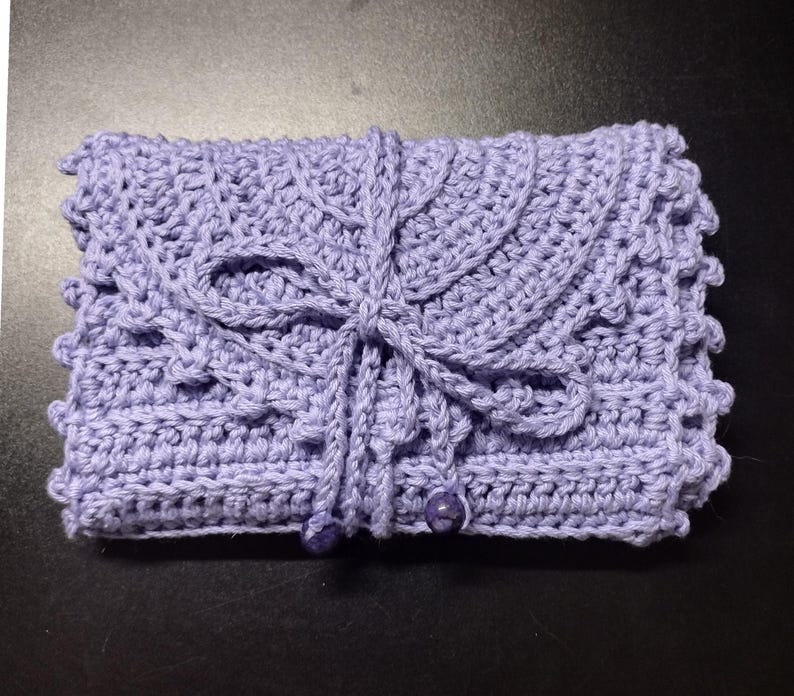 May include: A handmade, lavender crochet clutch. The clutch features a detailed, textured stitch pattern, a decorative flap, and a tie closure. Two small, round beads secure the closure. The overall design is intricate and delicate.