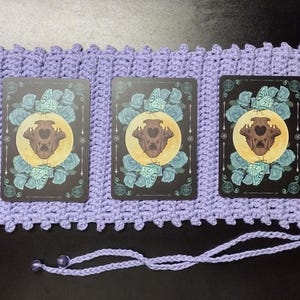 May include: A lavender crochet tarot card holder with a decorative edge and a drawstring closure. The holder contains a deck of tarot cards with a design featuring a skull, moon, and floral elements.