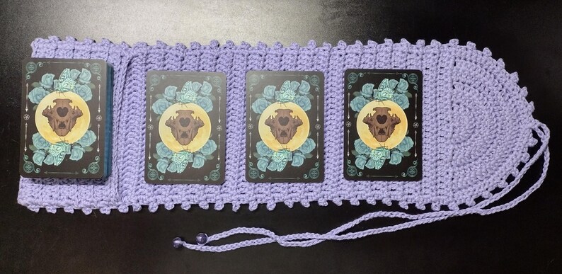 May include: A lavender crochet tarot card holder with a decorative edge and a drawstring closure. The holder contains a deck of tarot cards with a design featuring a skull, moon, and floral elements.