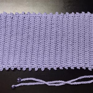 May include: A lavender crocheted phone sleeve with a drawstring closure. The sleeve has a textured, ribbed design and a scalloped edge. The drawstring is made of the same yarn and has two small, decorative beads at the ends.