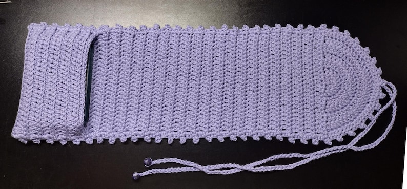 May include: A lavender crocheted phone sleeve with a drawstring closure. The sleeve has a textured, ribbed design and a scalloped edge. The drawstring is made of the same yarn and has two small, decorative beads at the ends.