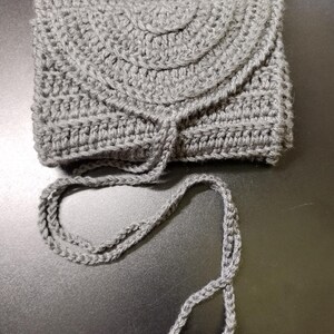 May include: A gray crocheted wallet with a flap closure and a long, braided drawstring. The wallet is rectangular with a rounded top and a textured surface. The drawstring ends with two small, round beads.
