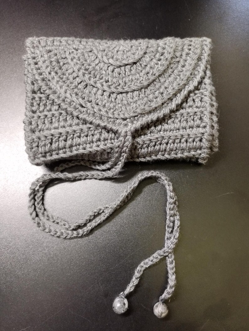 May include: A gray crocheted wallet with a flap closure and a long, braided drawstring. The wallet is rectangular with a rounded top and a textured surface. The drawstring ends with two small, round beads.