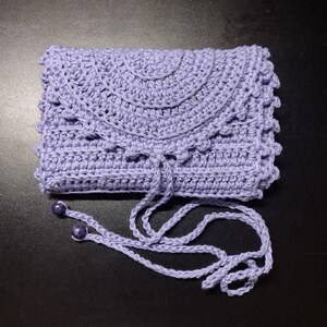 May include: A handmade, lavender crochet clutch with a flap closure and a drawstring. The clutch features a decorative scalloped edge and a matching drawstring with two purple beads. The bag is made of soft yarn and is suitable for carrying small items.