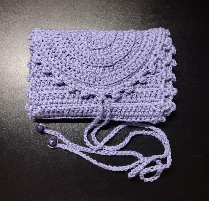 May include: A handmade, lavender crochet clutch with a flap closure and a drawstring. The clutch features a decorative scalloped edge and a matching drawstring with two purple beads. The bag is made of soft yarn and is suitable for carrying small items.
